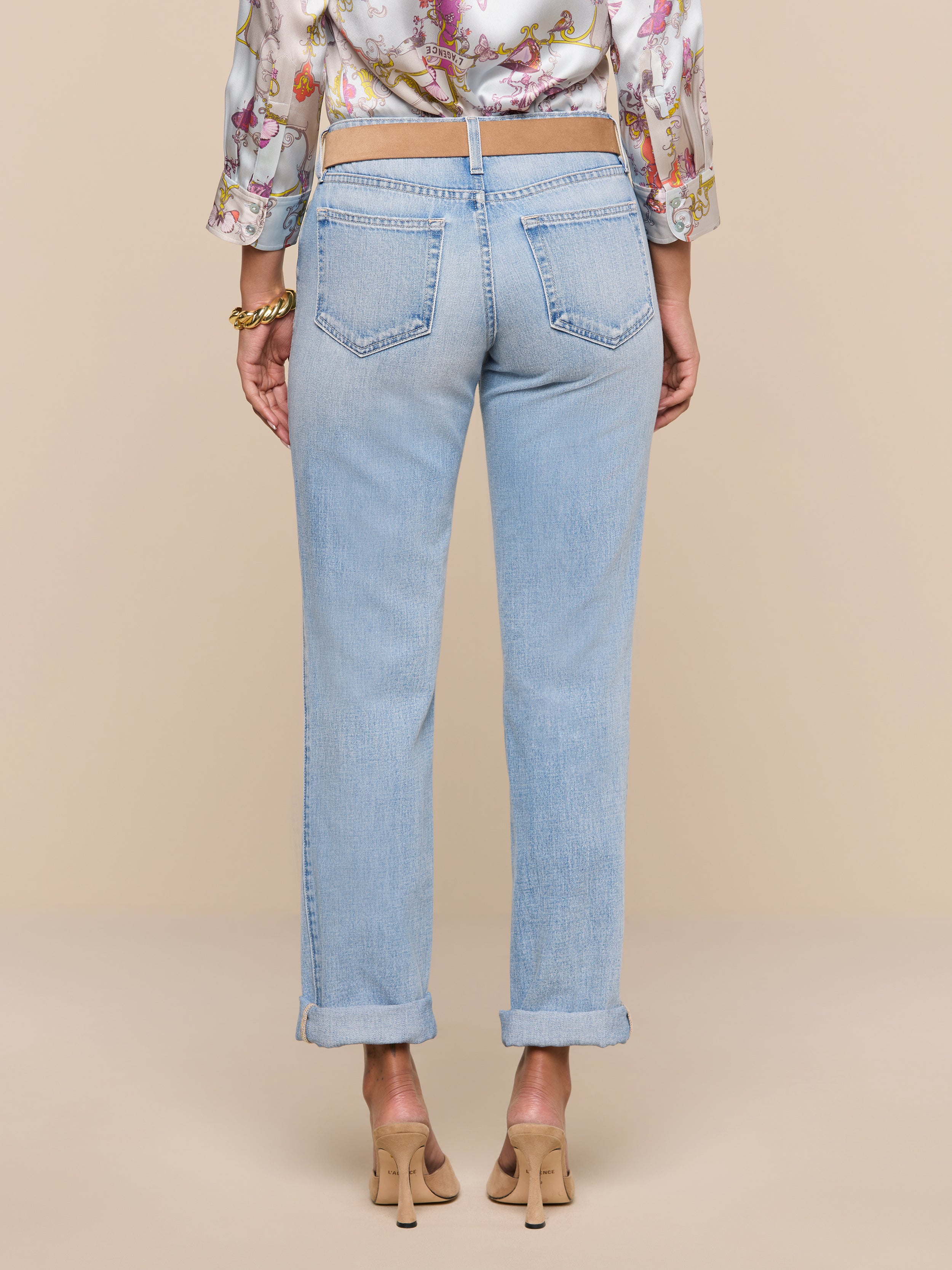 A person stands facing away against a plain white background, wearing the Ashby Low-Rise Slouch Jean in a vintage-inspired light wash, paired with a floral blouse and beige heels.