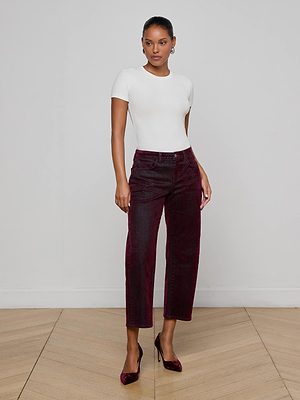 A person stands indoors against a plain wall, wearing a fitted white short-sleeve top, L'AGENCE Dolores Flocked Bow-Leg Jean in bordeaux velvet, and matching burgundy heels on a light wooden floor.