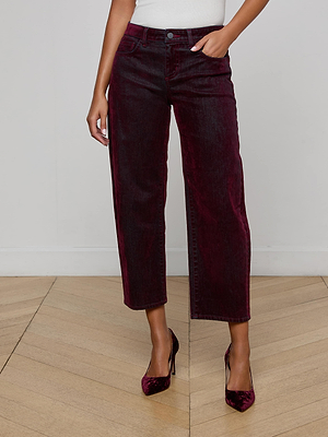 A person wears L'AGENCE Dolores Flocked Bow-Leg Jeans in dark bordeaux with a white top and matching pointed-toe heels, standing on a light wooden floor against a plain white wall.
