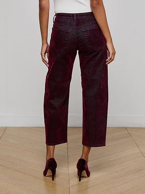 A person stands on a wooden floor facing away from the camera, wearing L'AGENCE Dolores Flocked Bow-Leg Jeans in bordeaux, a white top, and matching burgundy heeled shoes.