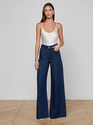 A woman with long brown hair and a neutral expression stands against a light-gray wall wearing the Pacey Wide-Leg Jean in dark wash, styled with a white satin camisole and white heeled sandals.