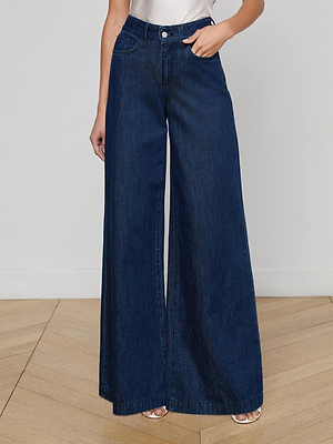 Someone stands on a light wooden floor wearing the Pacey Wide-Leg Jean in a dark wash with a white top, one hand in their pocket; their face is not visible.