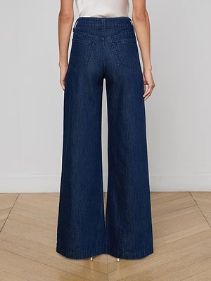 A person faces away, wearing the Pacey Wide-Leg Jean in a dark wash, featuring high-rise and two back pockets. They stand on a wooden floor with a plain, light-colored wall in the background.