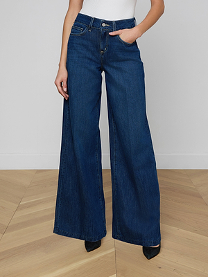 Wearing the Pacey Wide-Leg Jean in all-cotton denim with a white top, a person stands on a light wood floor with one hand in their pocket. The high-rise jeans drape over most of their black pointed-toe shoes.