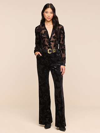 A woman stands against a beige background, wearing a sheer black blouse adorned with a delicate butterfly print. She pairs it with the Frances Bootcut Trouser—high-waisted black velvet pants featuring a chic bootcut silhouette and a matching belt. Her long dark hair is worn down, and she completes the look with classic black shoes.
