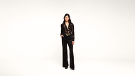A person stands in a studio against a plain white background, wearing a black patterned pantsuit with wide-leg trousers and black shoes, with one hand relaxed by their side and the other slightly raised.