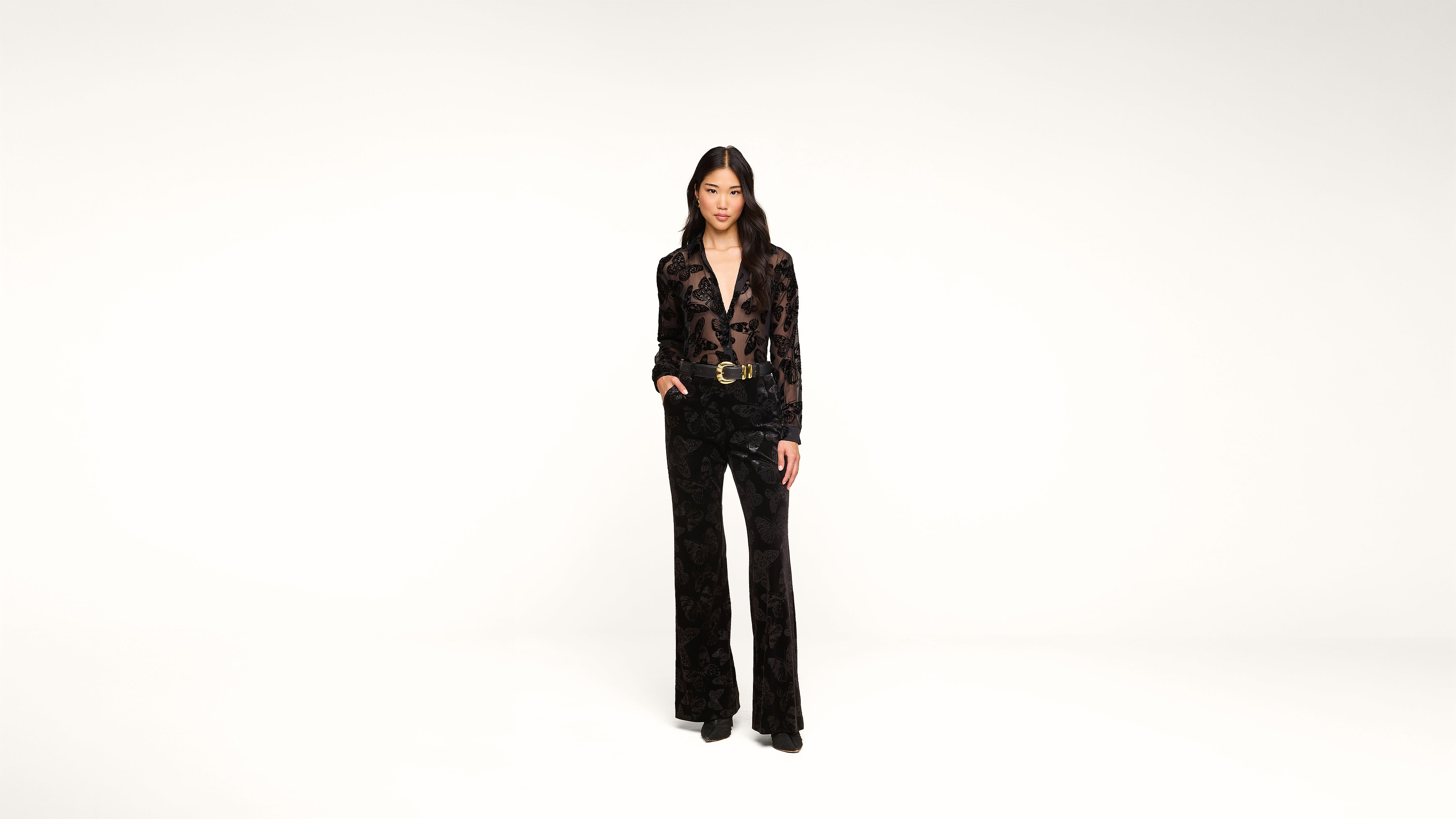 A person stands in a studio against a plain white background, wearing a black patterned pantsuit with wide-leg trousers and black shoes, with one hand relaxed by their side and the other slightly raised.