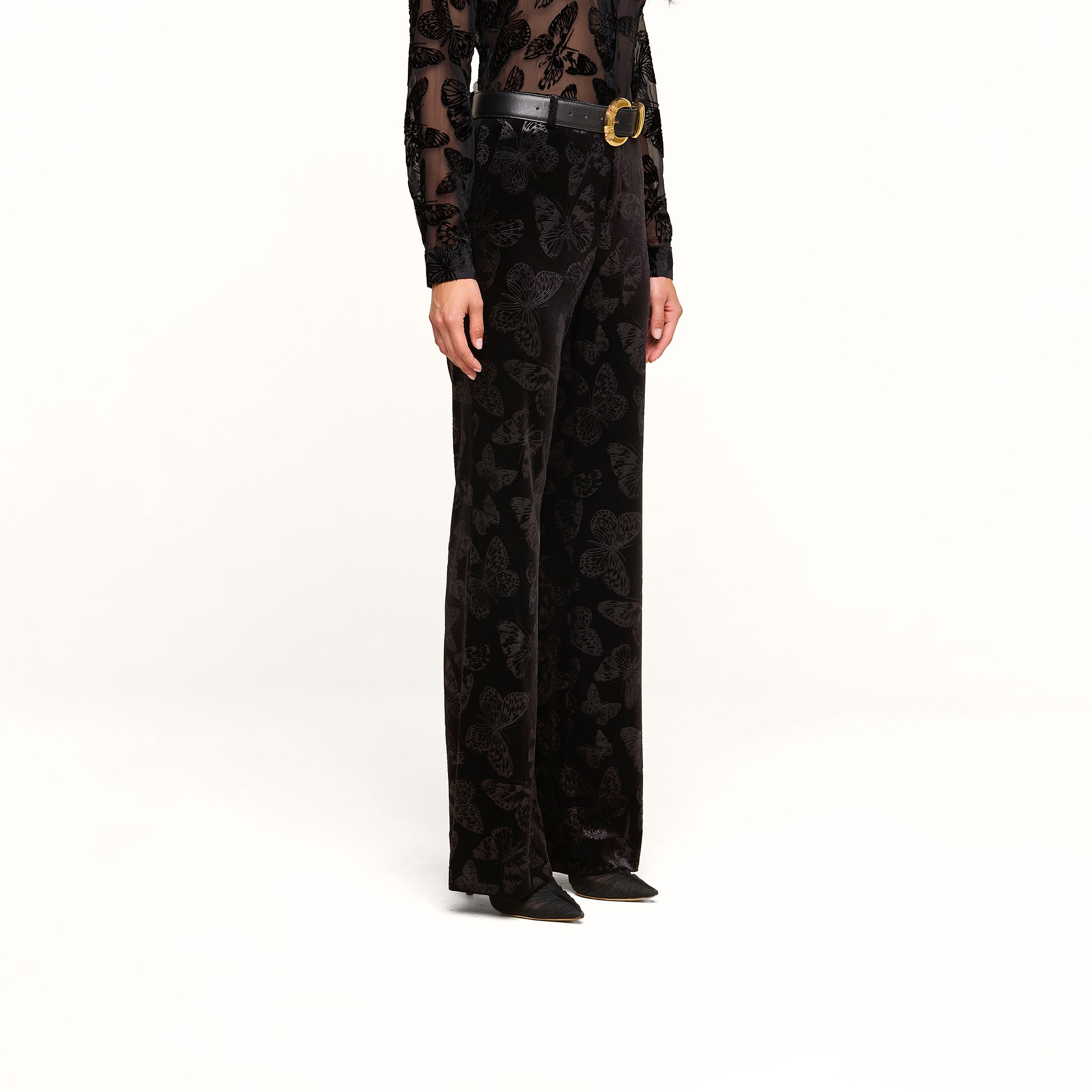 A person is shown from the waist down against a white background, wearing the Frances Bootcut Trouser in black stretch velvet with a subtle butterfly print. They have paired the high-waisted, wide-leg pants with black pointed-toe shoes and a sheer black top.