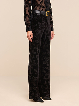 A person wears the Frances Bootcut Trouser in black, featuring a striking butterfly print, paired with a matching sheer black top, a black belt with a gold buckle, and pointed black shoes. The background is plain beige.