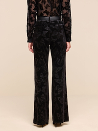 A person stands facing away, wearing the Frances Bootcut Trouser in black stretch velvet with a subtle butterfly print and a matching sheer top against a plain white background.