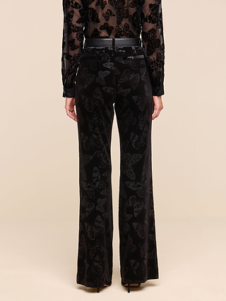 A person wearing the Frances Bootcut Trouser, featuring black velvet with a butterfly print and a bootcut silhouette, paired with a sheer black top, photographed from the back against a plain beige background.