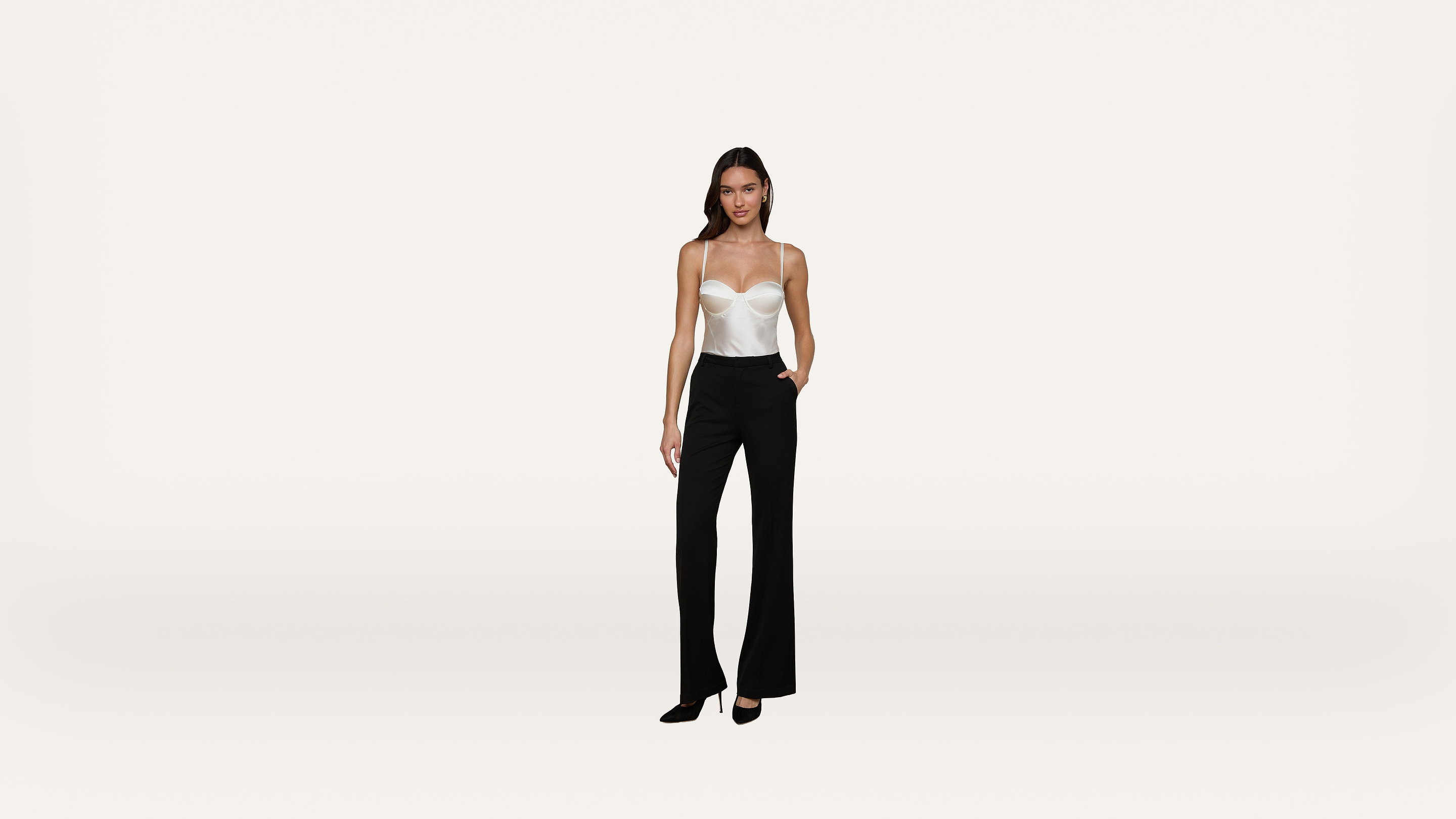 A woman stands against a plain white background, wearing a white strapless top, black high-waisted pants, and black heels.