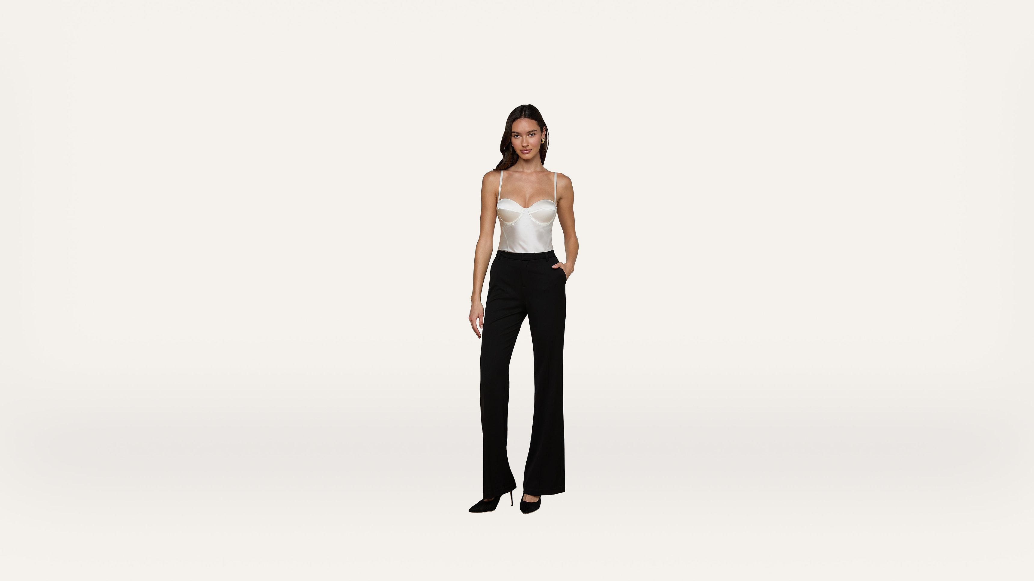 A woman stands against a plain white background, wearing a white strapless top, black high-waisted pants, and black heels.