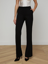 A woman stands against a plain background with one hand in her pocket, wearing black Frances Bootcut Trousers made from stretch suit fabric, a white sleeveless top, and black heels.