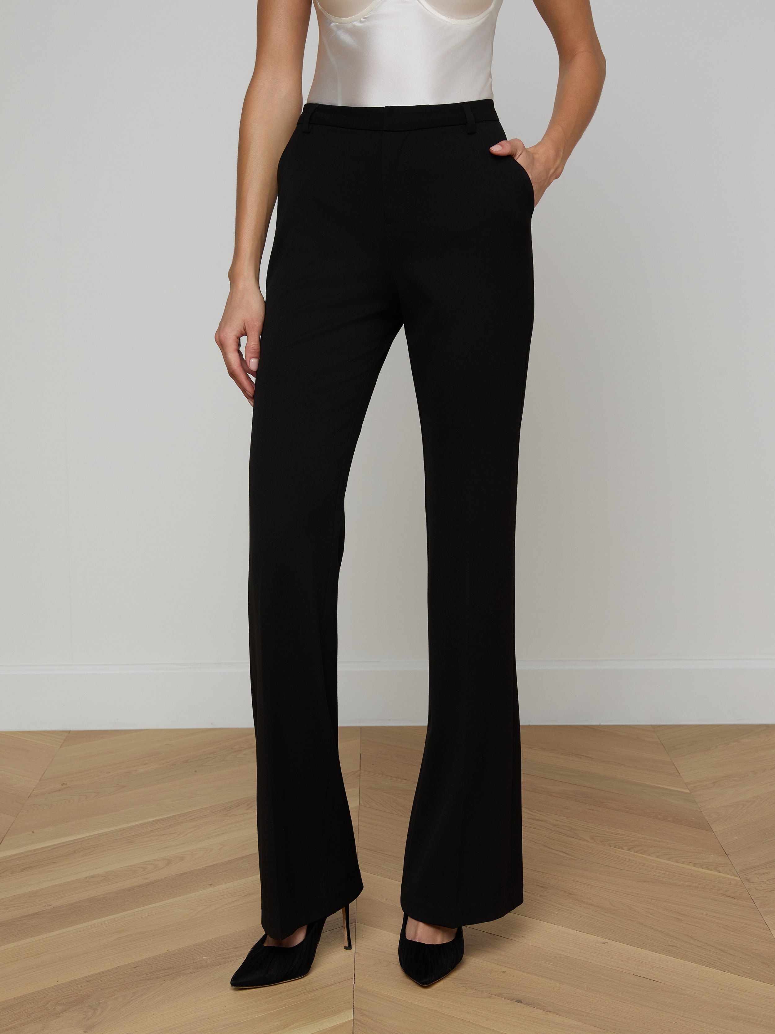 A woman stands against a plain background with one hand in her pocket, wearing black Frances Bootcut Trousers made from stretch suit fabric, a white sleeveless top, and black heels.