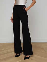 A woman wears the Frances Bootcut Trouser, crafted from stretch suit fabric in black, paired with a sleeveless top and black heels, standing with one hand in her pocket against a plain light background.