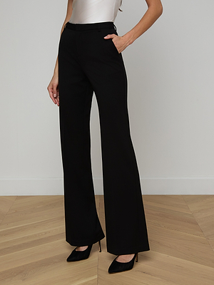 A person stands indoors on a wooden floor, wearing the Frances Bootcut Trouser—a high-waisted, wide-leg black pant in stretch suit fabric—styled with black pointed-toe heels and a tucked-in white top. Their left hand is in their pocket; their head is out of frame.