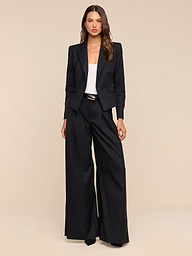 A woman stands against a beige background wearing the Chelsy Wide-Leg Pant in black pinstripe, paired with a matching blazer, a white top, and black shoes. She has long brown hair and a neutral expression, with her arms relaxed by her sides.