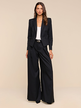 A woman stands against a beige background wearing the Chelsy Wide-Leg Pant in black pinstripe, paired with a matching blazer, a white top, and black shoes. She has long brown hair and a neutral expression, with her arms relaxed by her sides.