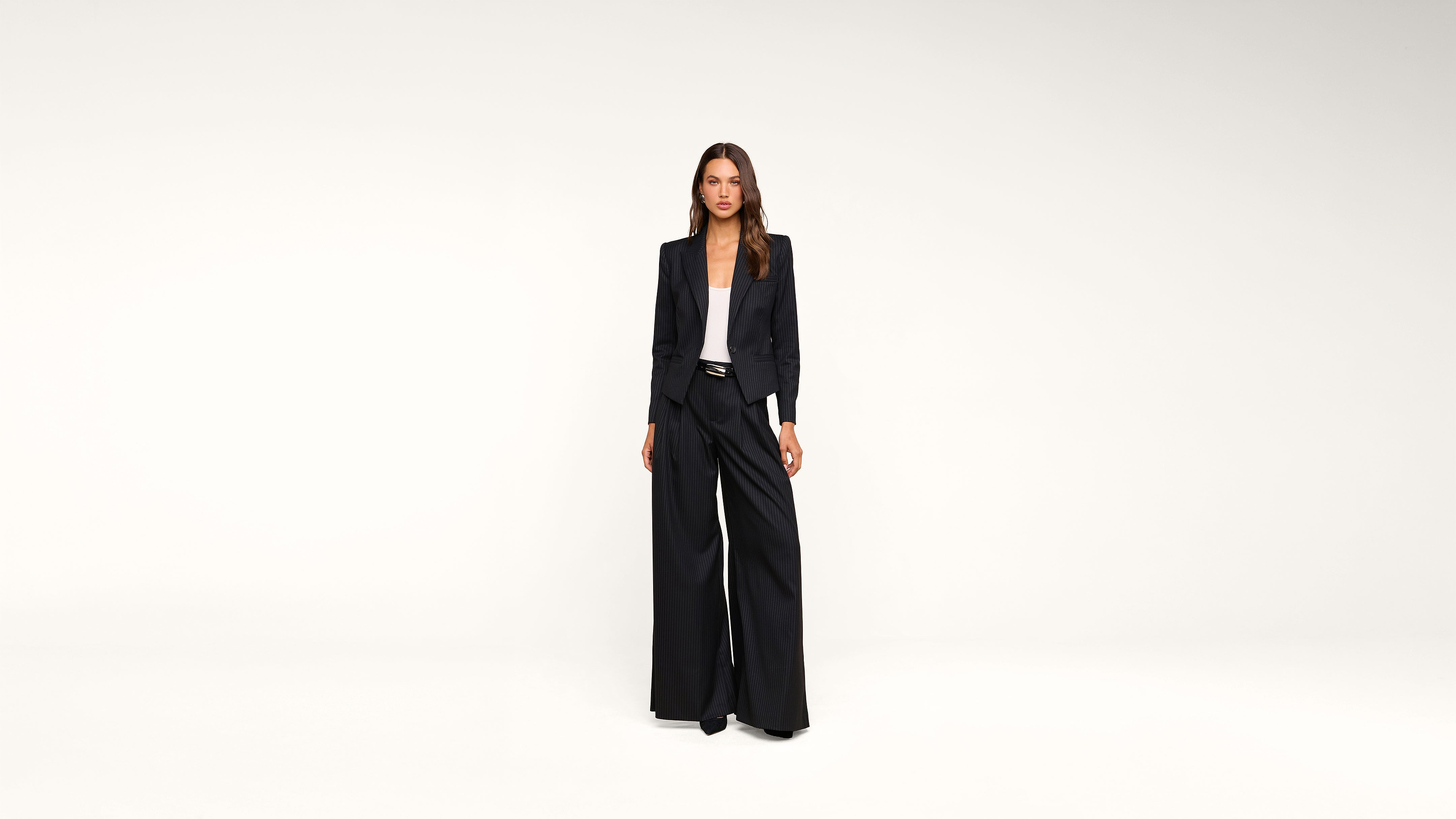 A woman stands against a plain white background, wearing a black blazer, wide-leg black trousers, and a white top. She faces forward with a neutral expression, hands by her sides.