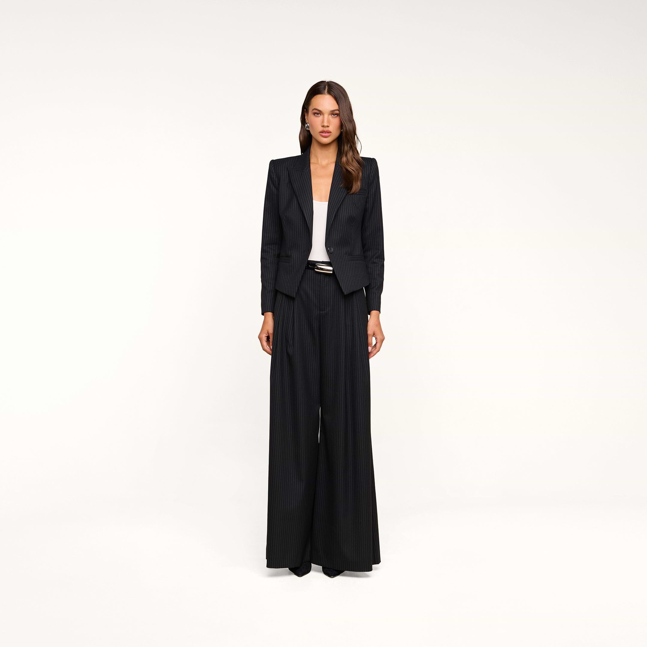 A woman stands against a plain white background wearing the Chelsy Wide-Leg Pant in black pinstripe, paired with a matching tailored blazer, white top, and black shoes. Her long brown hair is styled in loose waves, and she has a neutral facial expression.