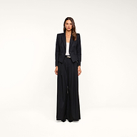 A woman stands facing forward against a plain beige background, wearing the Chelsy Wide-Leg Pant in black pinstripe, paired with a white top and black pointed shoes.