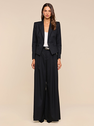 A woman stands facing forward against a plain beige background, wearing the Chelsy Wide-Leg Pant in black pinstripe, paired with a white top and black pointed shoes.