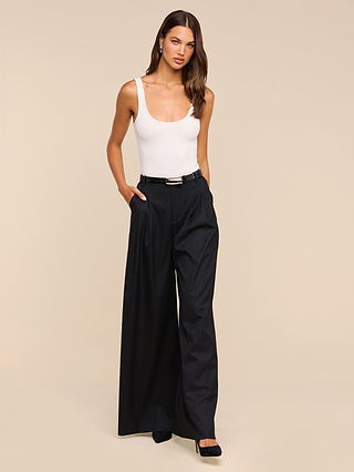 A woman stands against a plain beige background with her hands in her pockets, wearing the Chelsy Wide-Leg Pant—high-waisted and pinstriped—paired with a white tank top and black heels.