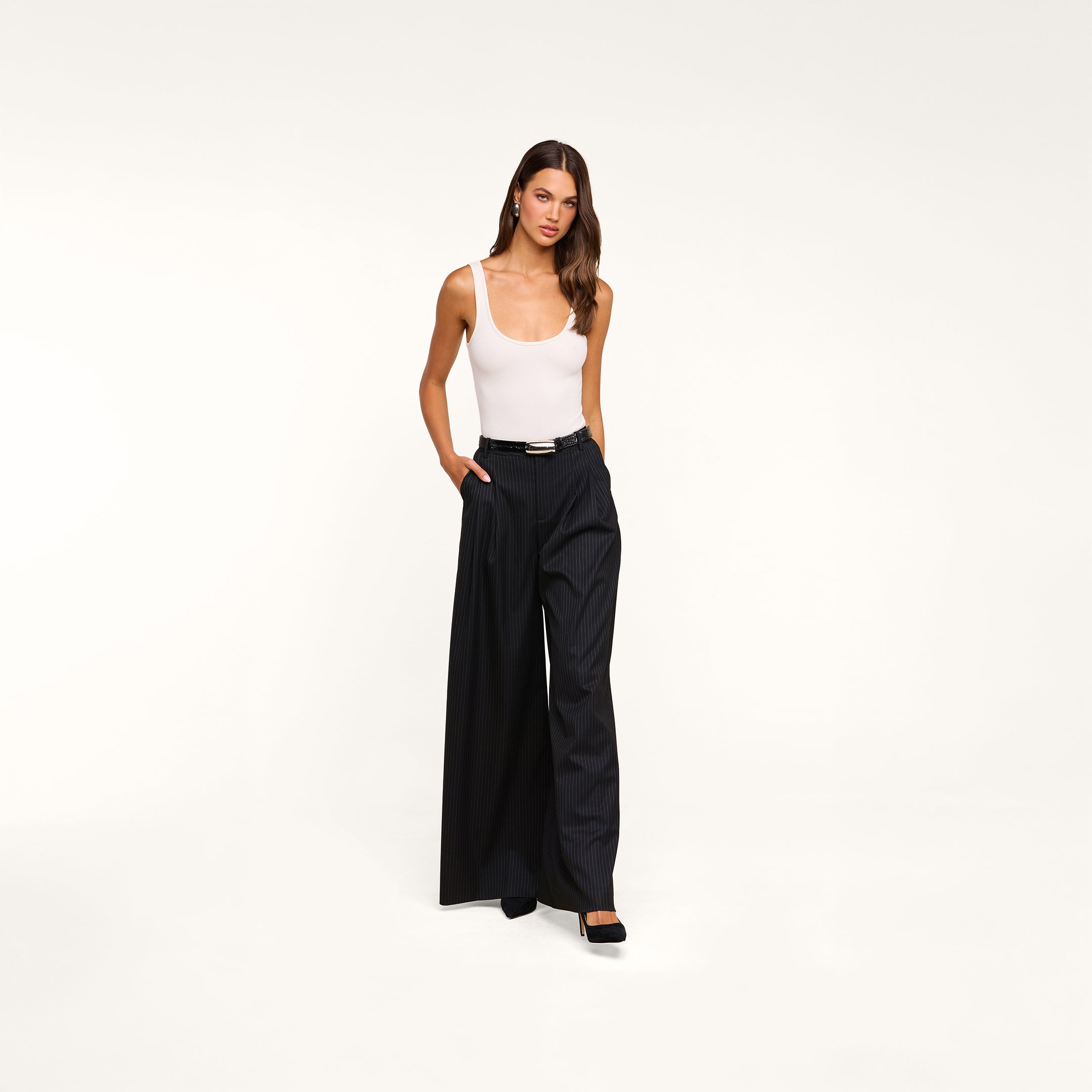 A woman stands against a plain beige background with her hands in her pockets, wearing the Chelsy Wide-Leg Pant—high-waisted and pinstriped—paired with a white tank top and black heels.
