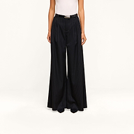 A person wearing a white sleeveless top and the Chelsy Wide-Leg Pant, featuring high-waisted pinstripes, styled with a black belt and pointed black shoes, stands against a plain beige background.