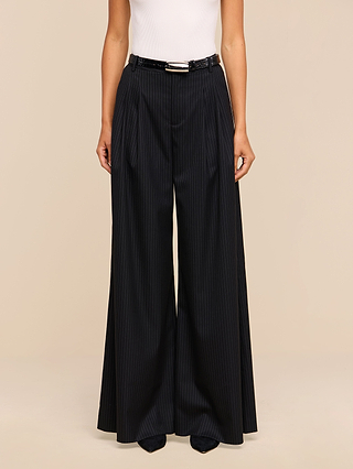 A person wearing a white sleeveless top and the Chelsy Wide-Leg Pant, featuring high-waisted pinstripes, styled with a black belt and pointed black shoes, stands against a plain beige background.