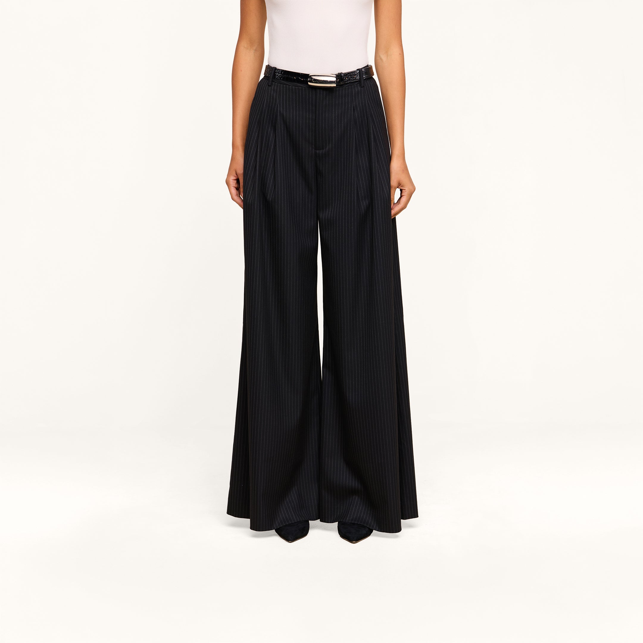 A person wearing a white sleeveless top and the Chelsy Wide-Leg Pant, featuring high-waisted pinstripes, styled with a black belt and pointed black shoes, stands against a plain beige background.