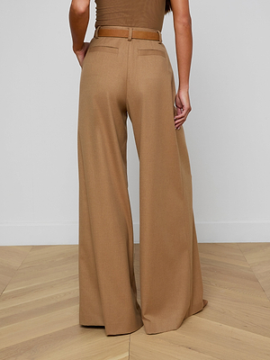 Someone stands facing away from the camera on a wooden floor against a plain white wall, wearing the Chelsy Wide-Leg Pant in tan suit fabric with a matching belt.