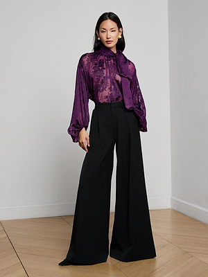 A woman poses confidently indoors against a light wall, wearing L'AGENCE's Chelsy Wide-Leg Pant paired with a purple sheer blouse featuring long, loose sleeves and a neck tie.