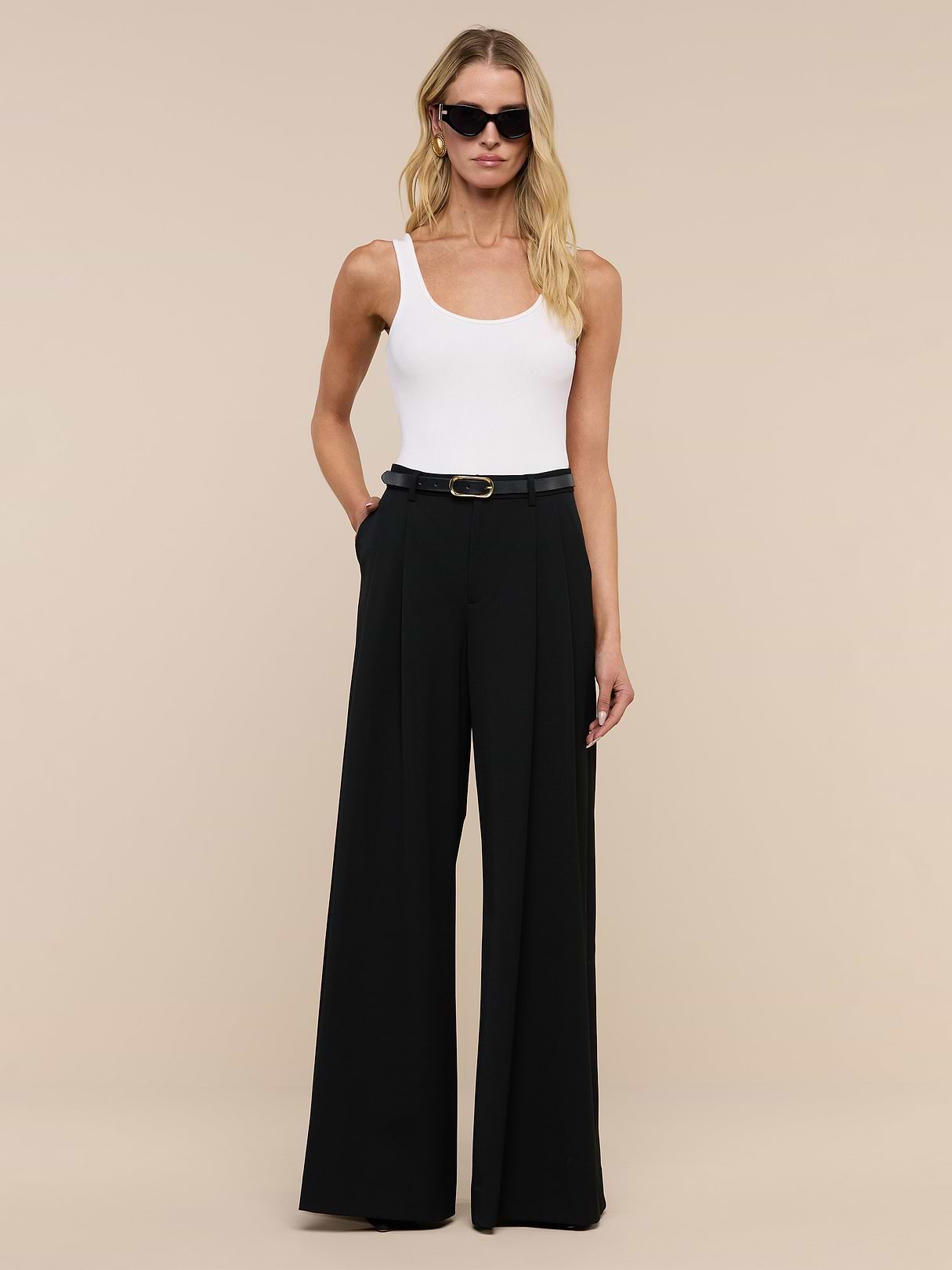 A woman in the Chelsy Wide-Leg Pant, a white sleeveless top, black belt, sunglasses, and gold earrings stands against a beige background with her hands in her pockets.