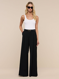 A woman wearing sunglasses, a white sleeveless top, and the Chelsy Wide-Leg Pant stands against a plain white background with her hands in her pockets.