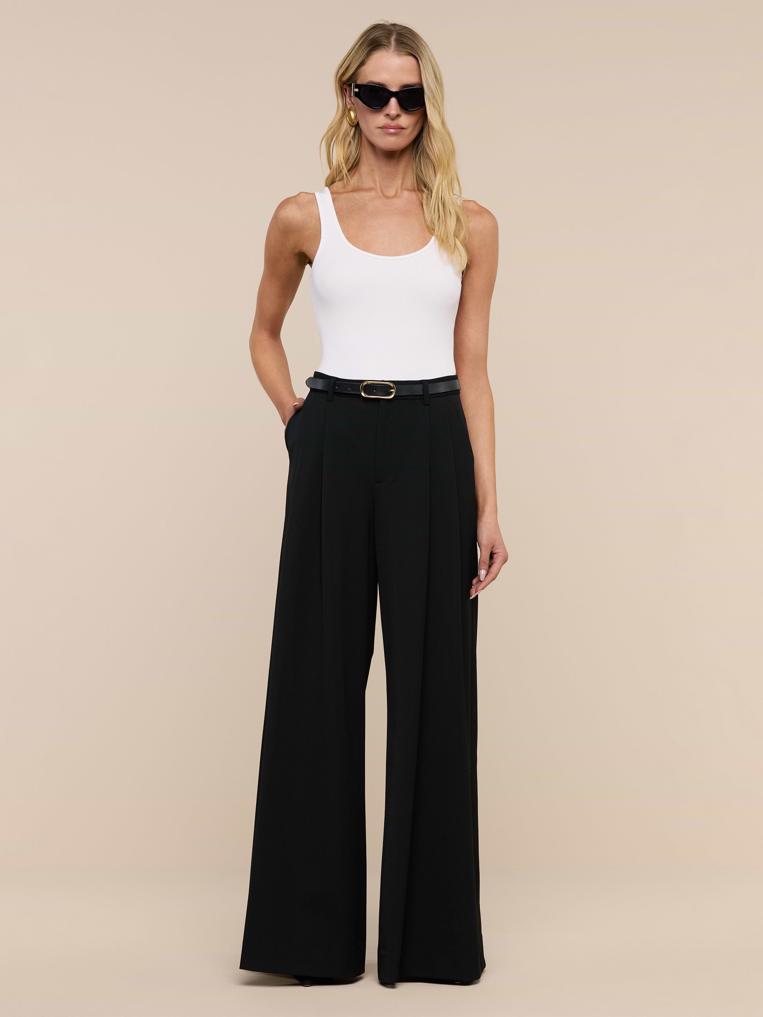 A woman wearing sunglasses, a white sleeveless top, and the Chelsy Wide-Leg Pant stands against a plain white background with her hands in her pockets.