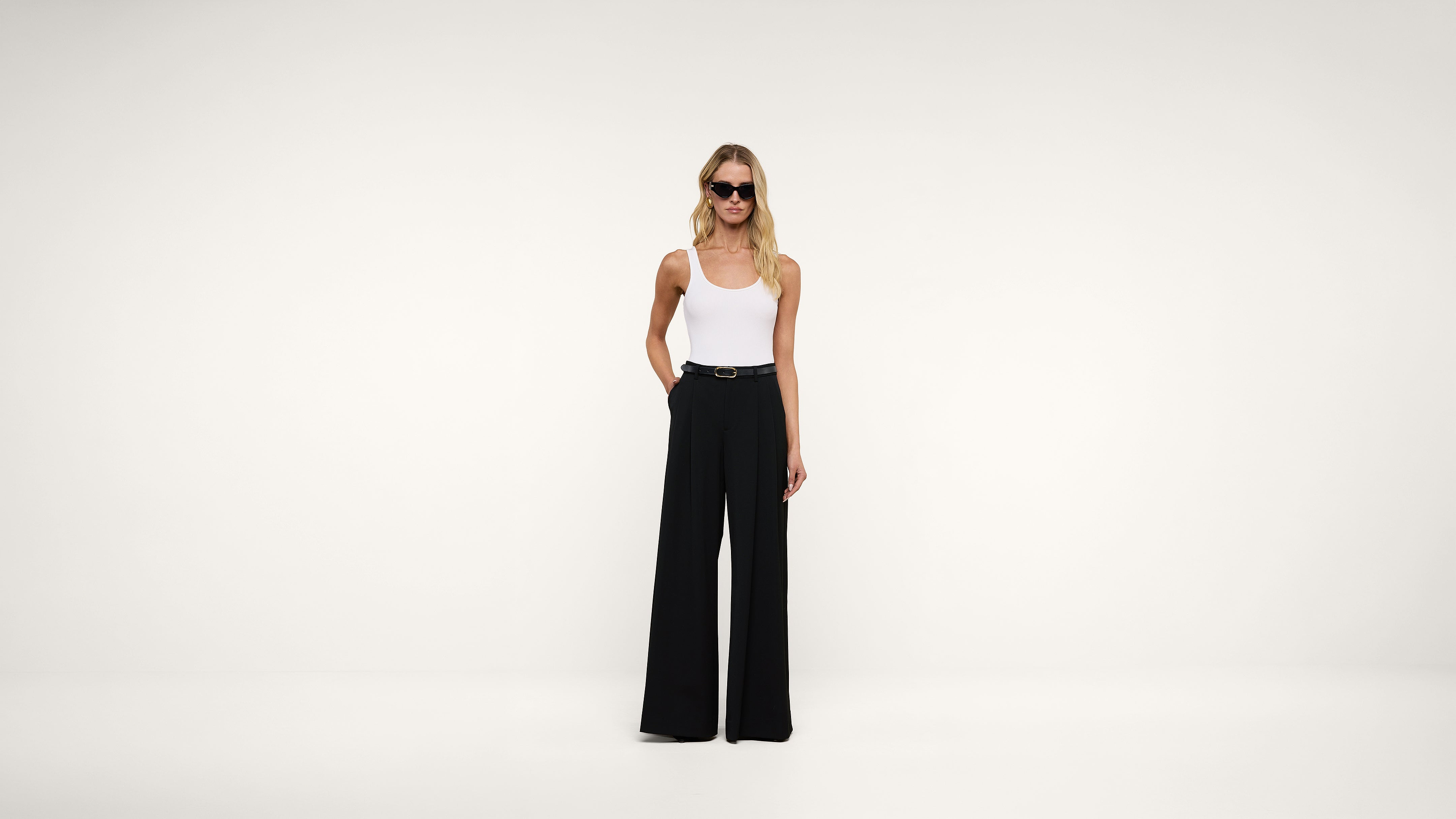 A woman wearing sunglasses, a white tank top, and wide-leg black pants stands against a plain white background, with one hand in her pocket.