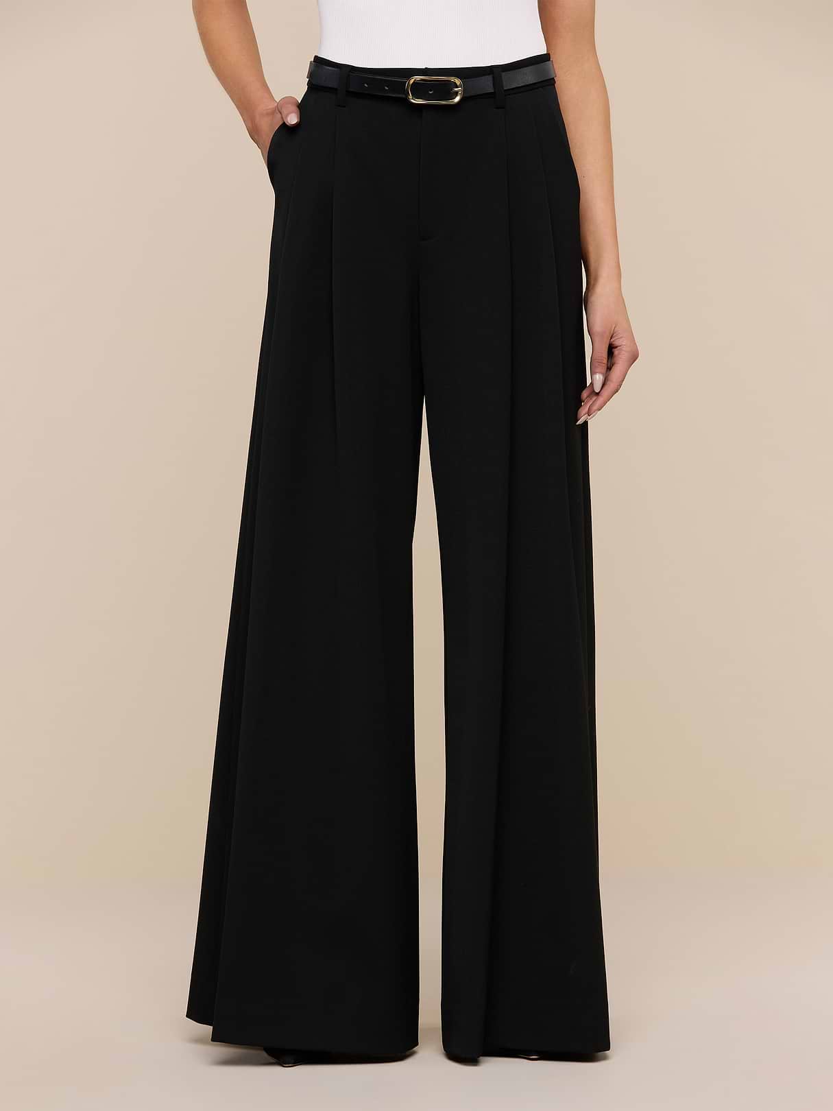 A person is wearing L'AGENCE Chelsy Wide-Leg Pant in black with a purple floral long-sleeve blouse, standing on a wooden floor against a plain white wall.