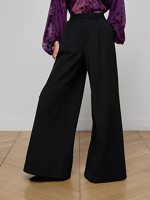 A person is wearing L'AGENCE Chelsy Wide-Leg Pant in black with a purple floral long-sleeve blouse, standing on a wooden floor against a plain white wall.