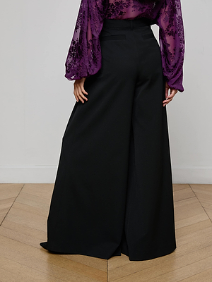 A person stands on a wooden floor wearing L'AGENCE Chelsy Wide-Leg Pant and a long-sleeve, sheer purple floral blouse; only the upper legs and torso are visible from the back.