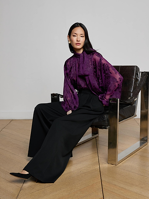 A woman with straight black hair sits calmly in a minimalist room, wearing a sheer purple floral blouse, black heels, and the L'AGENCE Chelsy Wide-Leg Pant.