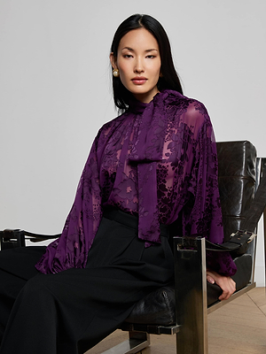 A woman with long dark hair sits on a black leather chair, wearing a sheer purple floral blouse with a bow and L'AGENCE Chelsy Wide-Leg Pant. She looks confidently at the camera.