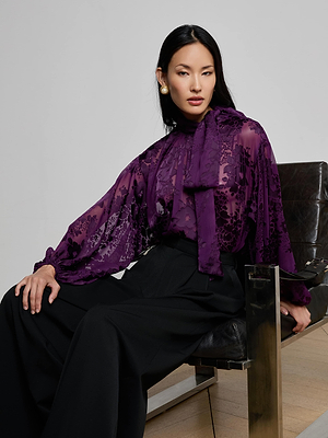A woman with long black hair sits on a modern chair, confidently looking at the camera. She wears a sheer purple floral blouse, large pearl earrings, and the L'AGENCE Chelsy Wide-Leg Pant.