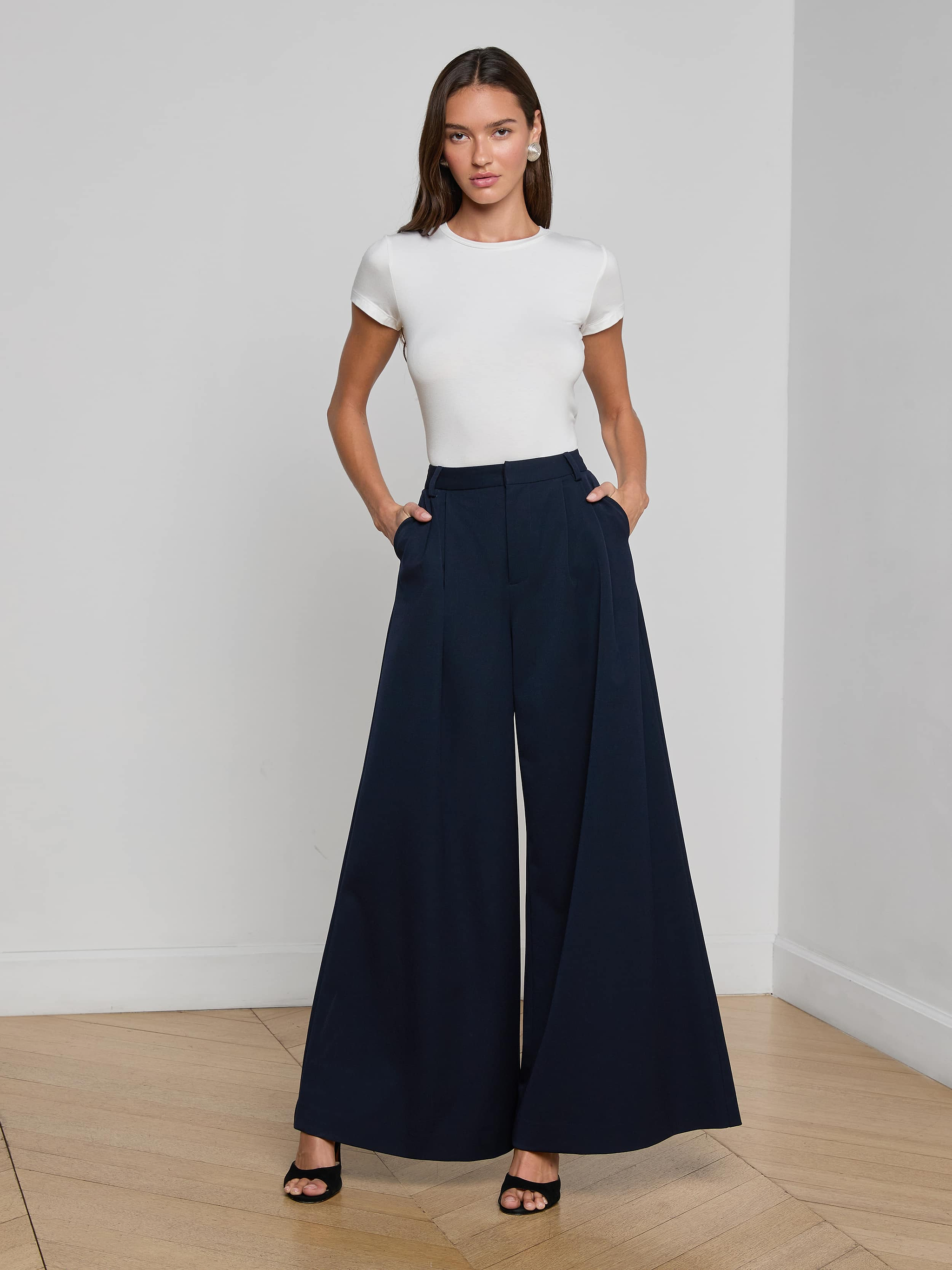A woman stands with her hands in her pockets, wearing the Chelsy Wide-Leg Pant—high-waisted trousers crafted from stretch suit fabric—paired with a fitted white short-sleeve top and black heeled sandals against a plain light background.