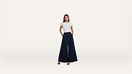 A woman stands against a white background, wearing a fitted white t-shirt, wide-leg navy blue pants, and black heeled sandals. She looks directly at the camera with her hands in her pockets.