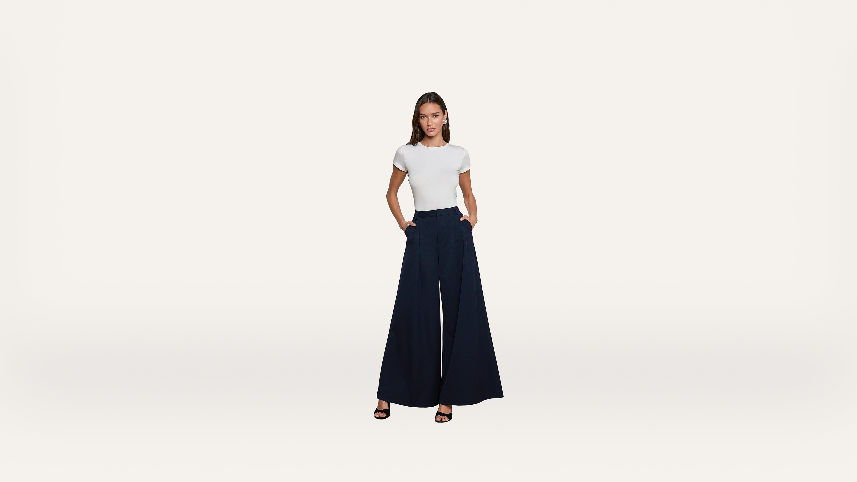 A woman stands against a white background, wearing a fitted white t-shirt, wide-leg navy blue pants, and black heeled sandals. She looks directly at the camera with her hands in her pockets.