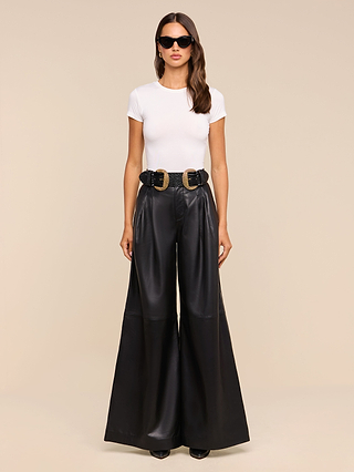 A woman stands facing forward against a plain beige background, wearing sunglasses, a white fitted t-shirt, and the Chelsy Leather Wide-Leg Pant in black styled with a black belt featuring two large gold buckles.
