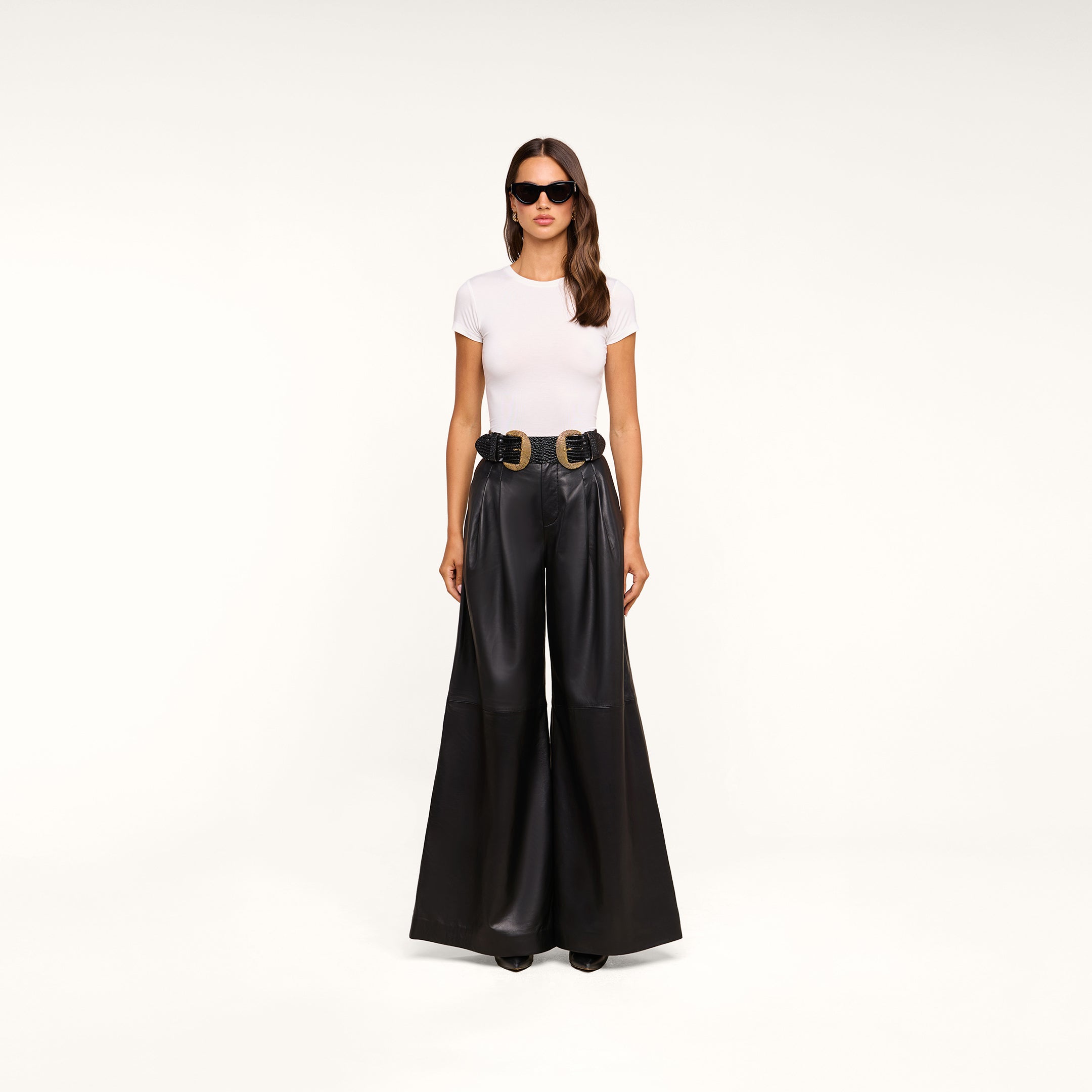 A woman wearing sunglasses, a white fitted t-shirt, and the Chelsy Leather Wide-Leg Pant with a double belt stands against a plain white background.