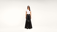 A woman wearing sunglasses, a white t-shirt, a wide black belt, and high-waisted, wide-leg black pants stands against a plain white background.